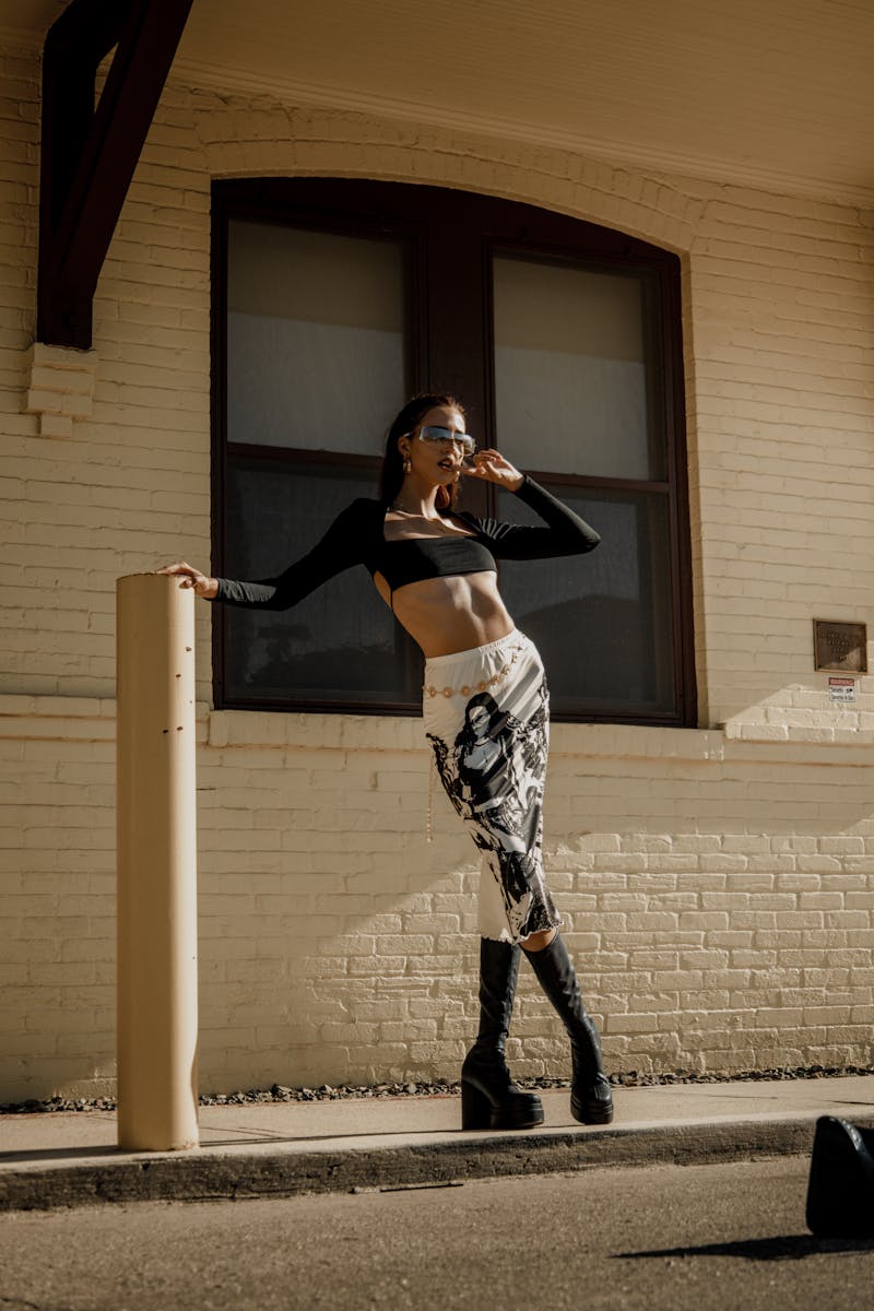 Fashionable woman in black top and skirt posing outdoors against a brick wall in daylight.